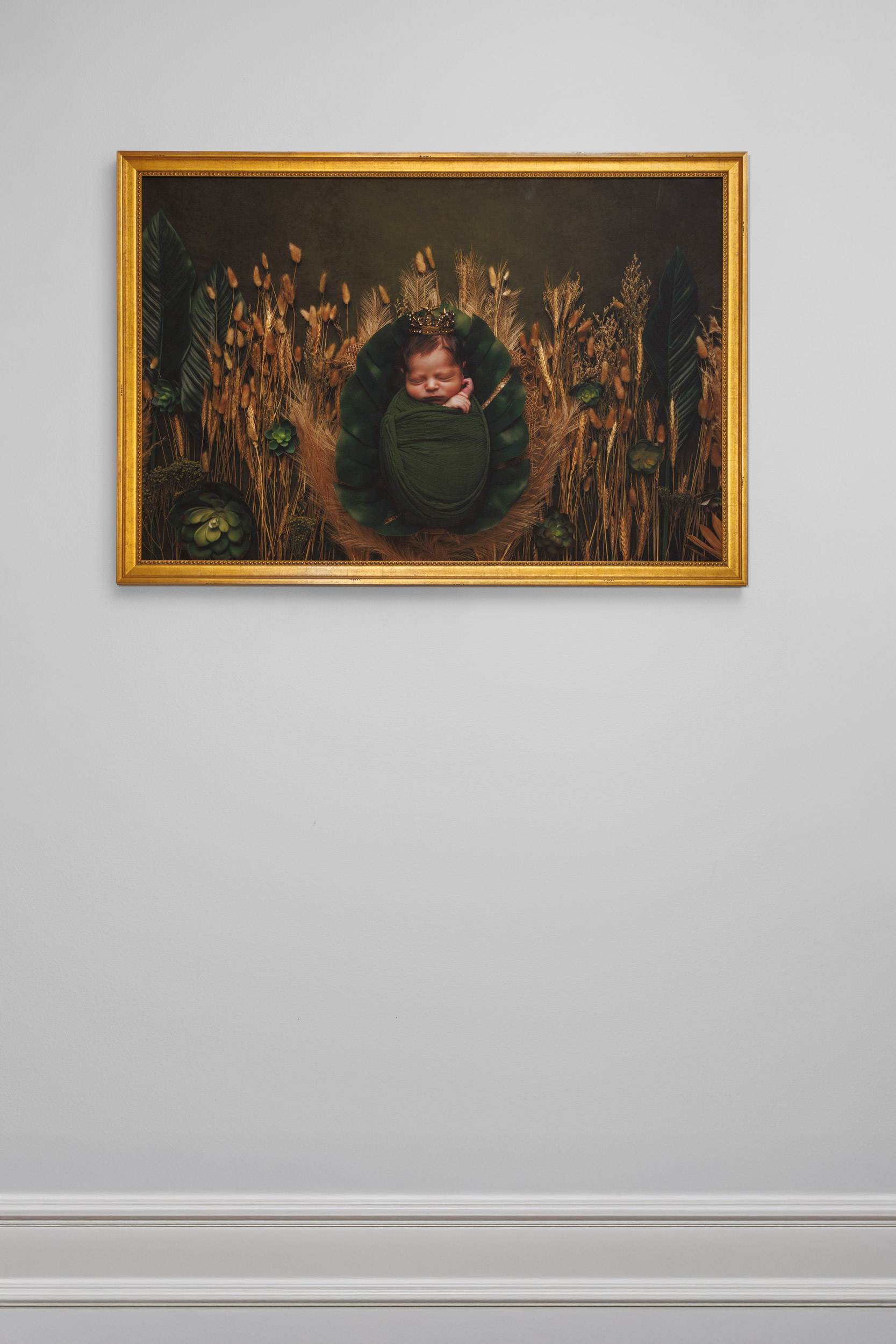 Fine art newborn portrait in an ornate gold frame displayed on a grey wall. The newborn is swaddled in deep green, nestled among golden wheat and dried botanicals, wearing a delicate gold crown. Fine art portrait photography by Anj Portrait Studio, Ankeny, Iowa, serving the greater Des Moines area.